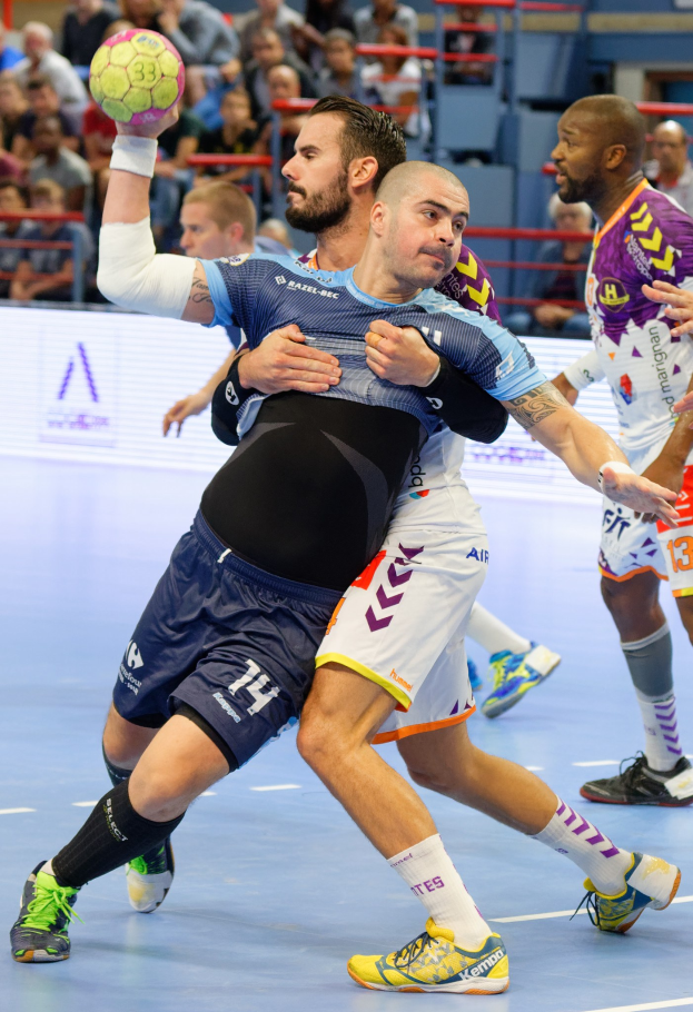 Männer spielen Handball in einem Stadion, einer hält den Ball, während andere im Hintergrund konkurrieren, mit Zuschauern auf Stühlen, Geländern und einer Wand im Hintergrund.