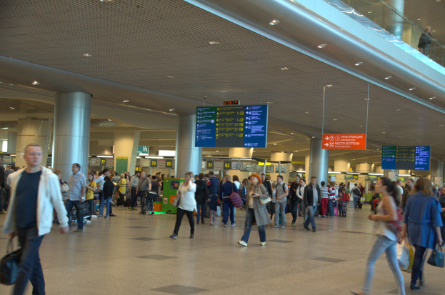 Eine große Gruppe von Menschen geht durch einen Flughafen, einige tragen Gepäck, mit Informationsschildern, Deckenleuchten, Säulen und einem Glasgeländer auf der rechten Seite.
