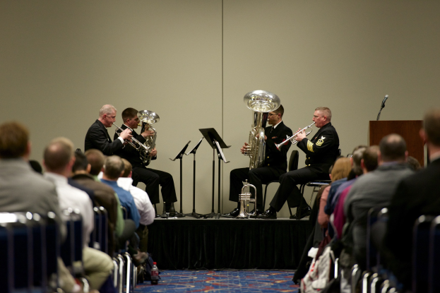Eine Gruppe von Männern, die auf einer Bühne Blasinstrumente spielen, mit Notenständern, einem Podium mit Mikrofon und einer Sitzreihe.