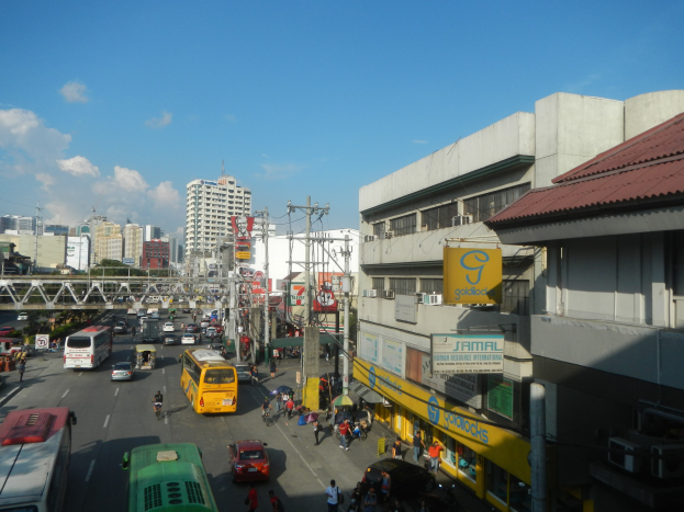 Eine belebte Stadtstraße mit Autos, Bussen, Fußgängern, Strommasten, Gebäuden mit Schildern, einer Brücke, Bäumen und einem bewölkten Himmel.
