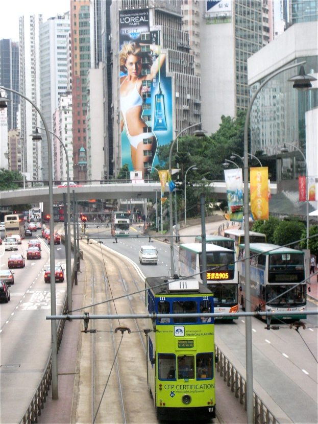 Eine belebte Stadtstra├če mit Doppeldeckerbussen, Autos und anderen Fahrzeugen, mit Pfosten, Laternen, Schildern, B├Ąumen, einer ├Überquerung, hohen Geb├Ąuden und einem klaren blauen Himmel, mit Text unten.