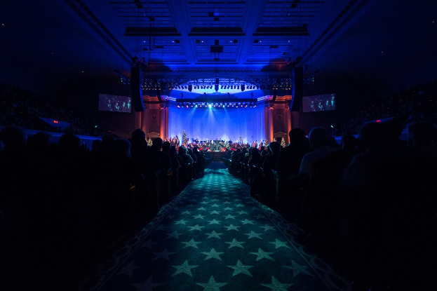 Ein großer Saal mit sitzenden Zuschauern, eine Bühne mit Musikern und Deckenleuchten und -fernseher, was auf ein Konzerthaus während der Nationalkonvention 2019 hinweist.