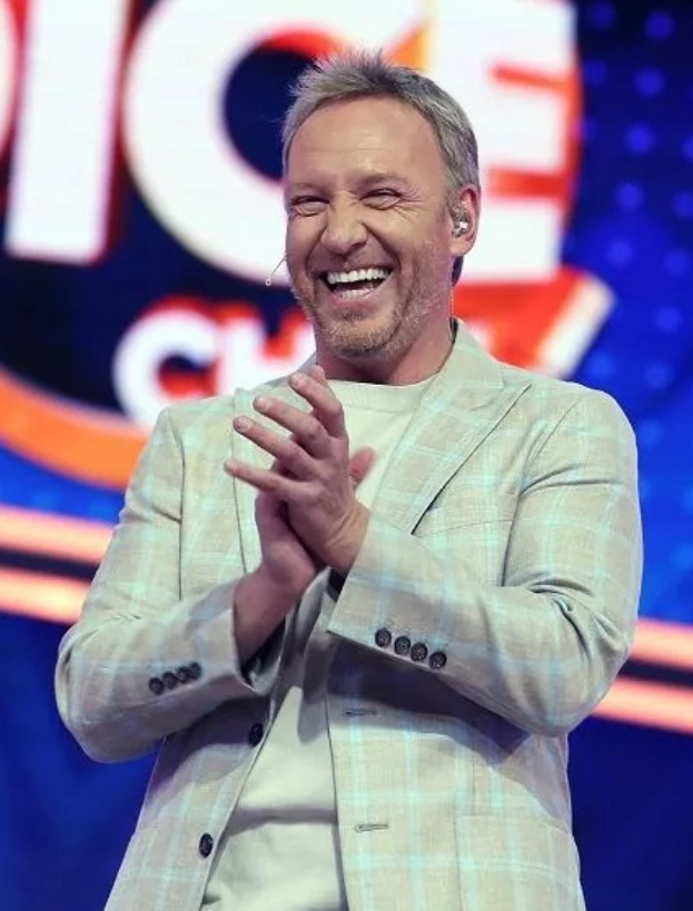 Ein Mann in einem Anzug, der auf einer Bühne klatscht, mit 'The Voice' auf einer Tafel im Hintergrund.