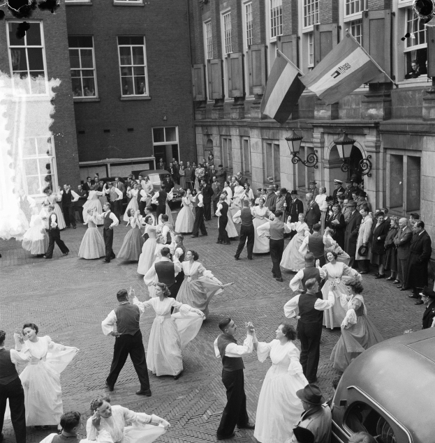 Eine Gruppe von Menschen, die auf der Straße vor einem Gebäude tanzen, mit einem geparkten Auto auf der rechten Seite und Fahnen und Lichtern an der Wand des Gebäudes im Hintergrund, dargestellt in Schwarz-Weiß.