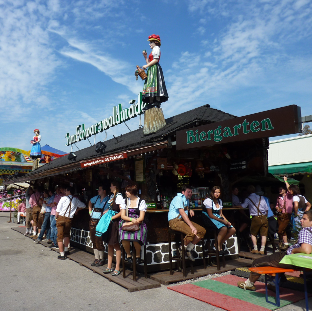 Eine Gruppe von Menschen vor einem Gebäude auf dem Oktoberfest in München, einige in traditioneller bayrischer Kleidung, andere auf Hockern sitzend mit Tischen voller Flaschen und einer Tafel im Hintergrund, Bäume und bewölkter Himmel sichtbar.