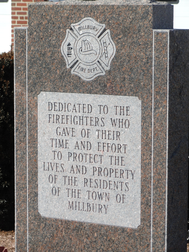 Gedenkstein mit einer Tafel für Feuerwehrleute in Millbury, vor einem Backsteingebäude mit umliegenden Pflanzen und Bäumen gesetzt.