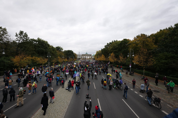 Eine große Gruppe von Menschen marschiert auf einer von Bäumen gesäumten Straße mit Laternenmasten, die Kameras halten, mit einem Gebäude und einem klaren Himmel im Hintergrund.