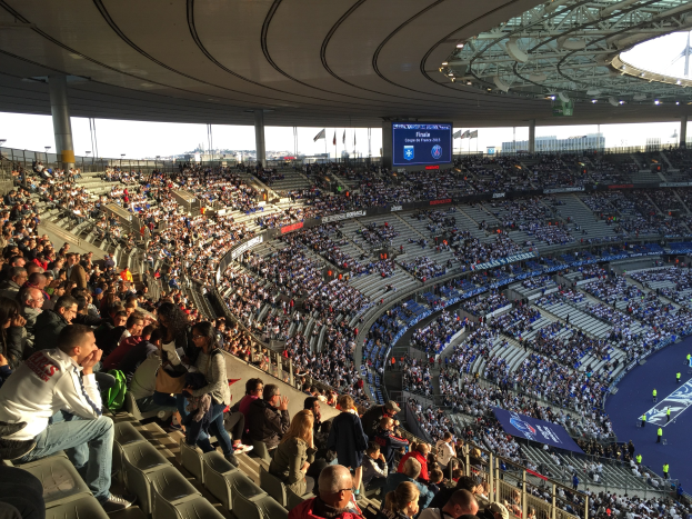 Eine große Menge Menschen in einem Stadion schaut ein Fußballspiel zu, mit einer Bühne, Fahnen, Stangen und einem Bildschirm im Hintergrund, und die Allianz Arena in München, Deutschland ist erkennbar.