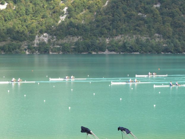 Eine Gruppe von Menschen bei einem Ruderwettbewerb auf einem Gewässer mit Bäumen und einem Hügel im Hintergrund, sowie Kleidung über Ständen am Boden.