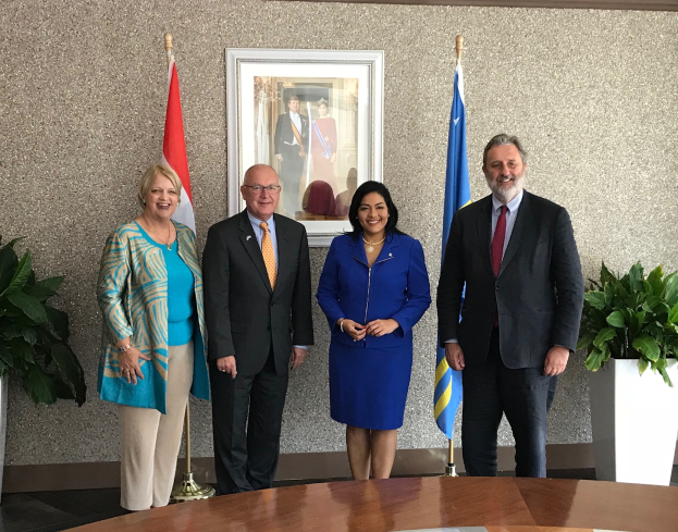 Eine Gruppe strahlender Menschen um einen Tisch mit Papieren versammelt, umgeben von Fahnen, Topfpflanzen und einem Foto auf dem Wand, feiert die Unterzeichnung eines Memorandums zwischen den Vereinigten Staaten und Kanada.