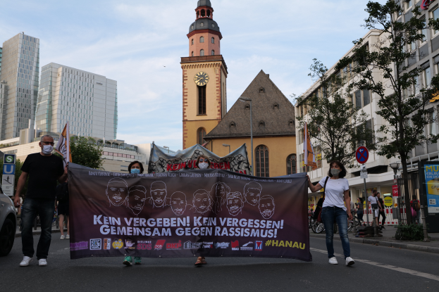 Eine Gruppe maskierter Personen marschiert auf einer Straße und hält ein Banner hoch, mit einem geparkten Auto auf der linken Seite, Gebäuden, Bäumen, Schildern, Pfählen und einem Uhrenturm im Hintergrund unter einem klaren blauen Himmel.