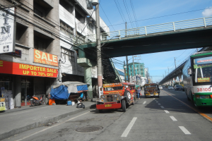 Eine belebte Stadtstraße mit Fahrzeugen, Gebäuden, Strommasten mit Drähten, einer Brücke und einem wolkenverhangenen Himmel im Hintergrund.