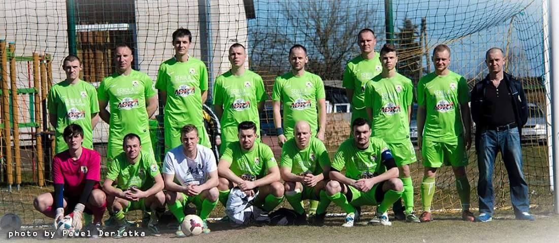Eine Gruppe von Männern in grünen T-Shirts steht auf einem Fußballfeld, einige in Hockstellung, mit einem Ball davor und einem Torpfosten rechts daneben, vor einer Kulisse aus Bäumen, Gebäuden und Fahrzeugen.