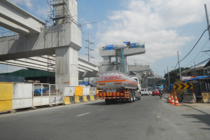 Ein großer Lkw fährt auf einer Straße neben einer Brücke, umgeben von Absperrkegeln, Strommasten mit Drähten, Gebäuden, Bäumen und einem bewölkten Himmel.