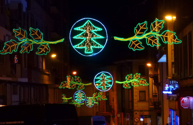 Eine nächtliche Stadtstraße, geschmückt mit festlichen Weihnachtslichtern in Form von Blättern, mit fahrenden Fahrzeugen und Gebäuden auf beiden Seiten.