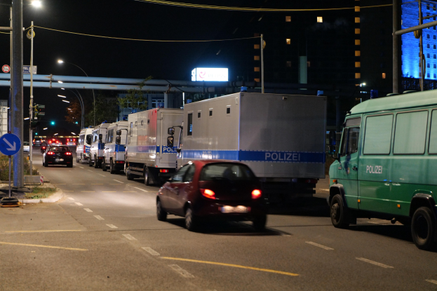 Lange Reihe von Polizeiwagen, die entlang einer Straße bei Nacht geparkt sind, mit Verkehr, Straßenschildern, Versorgungsmasten, Gebäuden und einer Brücke im Hintergrund.