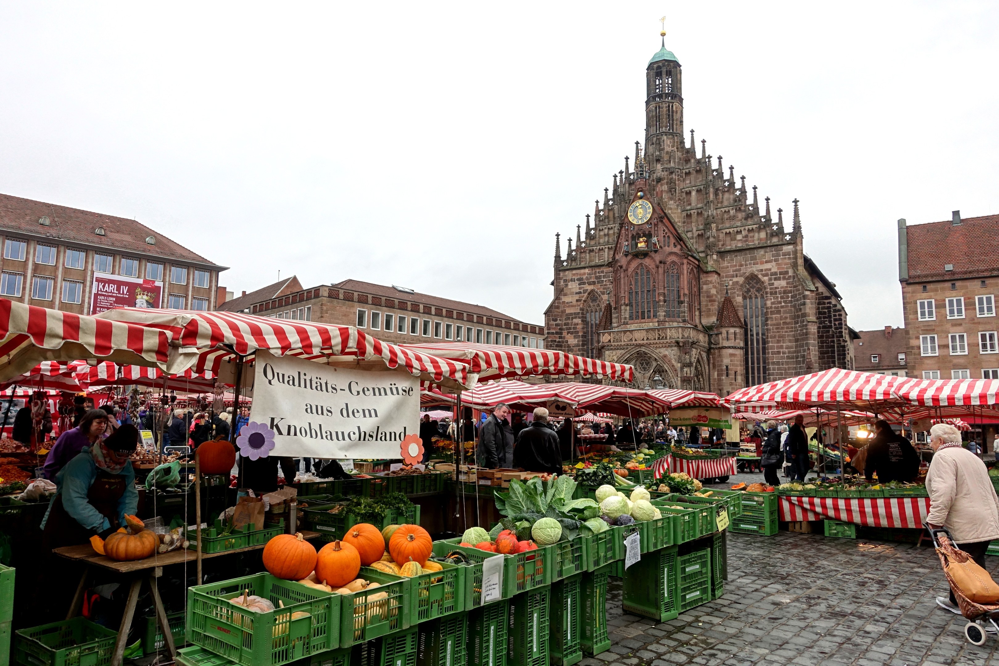 Ein belebter Markt in Nürnberg, Deutschland, mit Ständen voller Obst und Gemüse, Menschen mit Taschen und aufgestellten Zelten, sowie Gebäuden und einem Uhrenturm im Hintergrund unter einem sichtbaren Himmel.