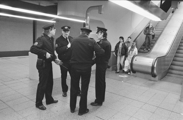 Schwarzes und weißes Bild von Polizeibeamten, die in einer U-Bahn-Station neben einer Rolltreppe mit ein paar Menschen darauf stehen.