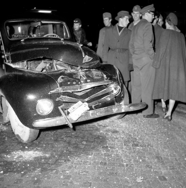 Ein Schwarz-Weiß-Bild eines verbeulten Autos am Straßenrand mit zersplitterten Fenstern und einer besorgten Menschenmenge drumherum.