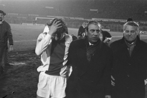 Eine Gruppe Männer in Not steht zusammen auf einem Fußballfeld, im Hintergrund ein Stadion, in einem Schwarz-Weiß-Bild.