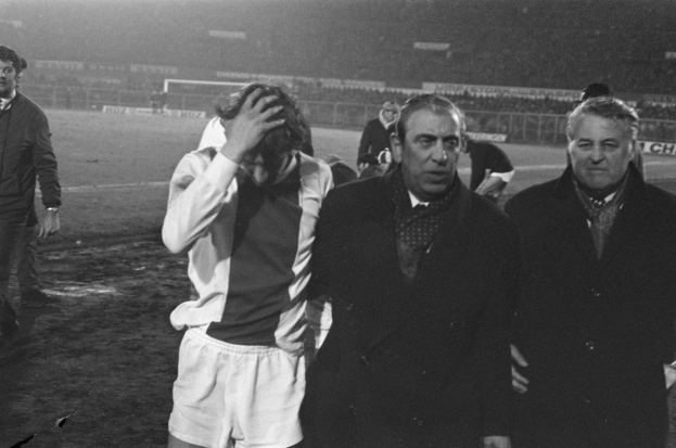 Eine Gruppe Männer in Not steht zusammen auf einem Fußballfeld, im Hintergrund ein Stadion, in einem Schwarz-Weiß-Bild.