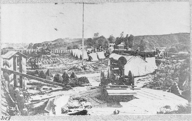 Schwarz-weißes Foto einer Baustelle mit einer Maschine im Vordergrund, umgeben von Hütten, Bäumen, Hügeln und Himmel, mit Text unten: "Die Bauarbeiten an einer neuen Brücke in der Stadt San Francisco, Kalifornien."