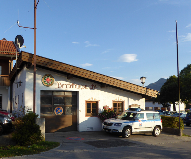 Polizeiauto vor einem Gebäude mit Fenstern und Türen geparkt, umgeben von Grünflächen einschließlich blühender Pflanzen, Gras und Bäumen, mit sichtbaren Straßeninfrastrukturen wie Masten, eine Schüsselantenne und ein Schild unter einem bewölkten Himmel.