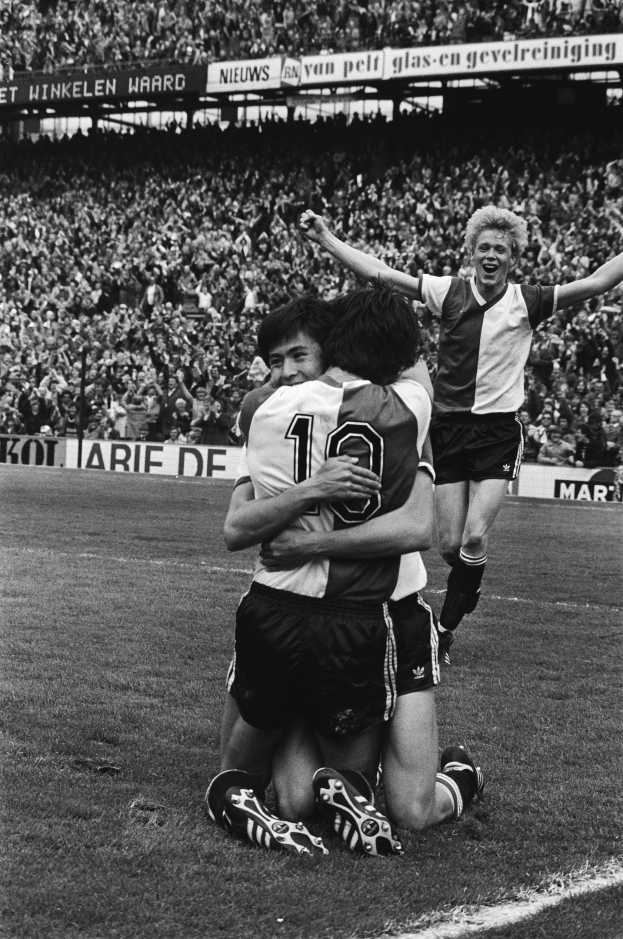 Drei Fussballspieler umarmen sich auf dem Boden beim Feiern eines Tors, mit jubelnder Menge und schwenkenden Fahnen im schwarz-weißen Hintergrund.