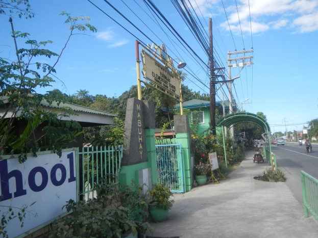 Einfahrt zu einer Schule mit einem "Schule"-Schild, Fahrzeuge auf der Straße, ein Torbogen, ein Tor, eine Namensschild, Pflanzen, Bäume, Strommasten mit Drähten und ein bewölkter Himmel.