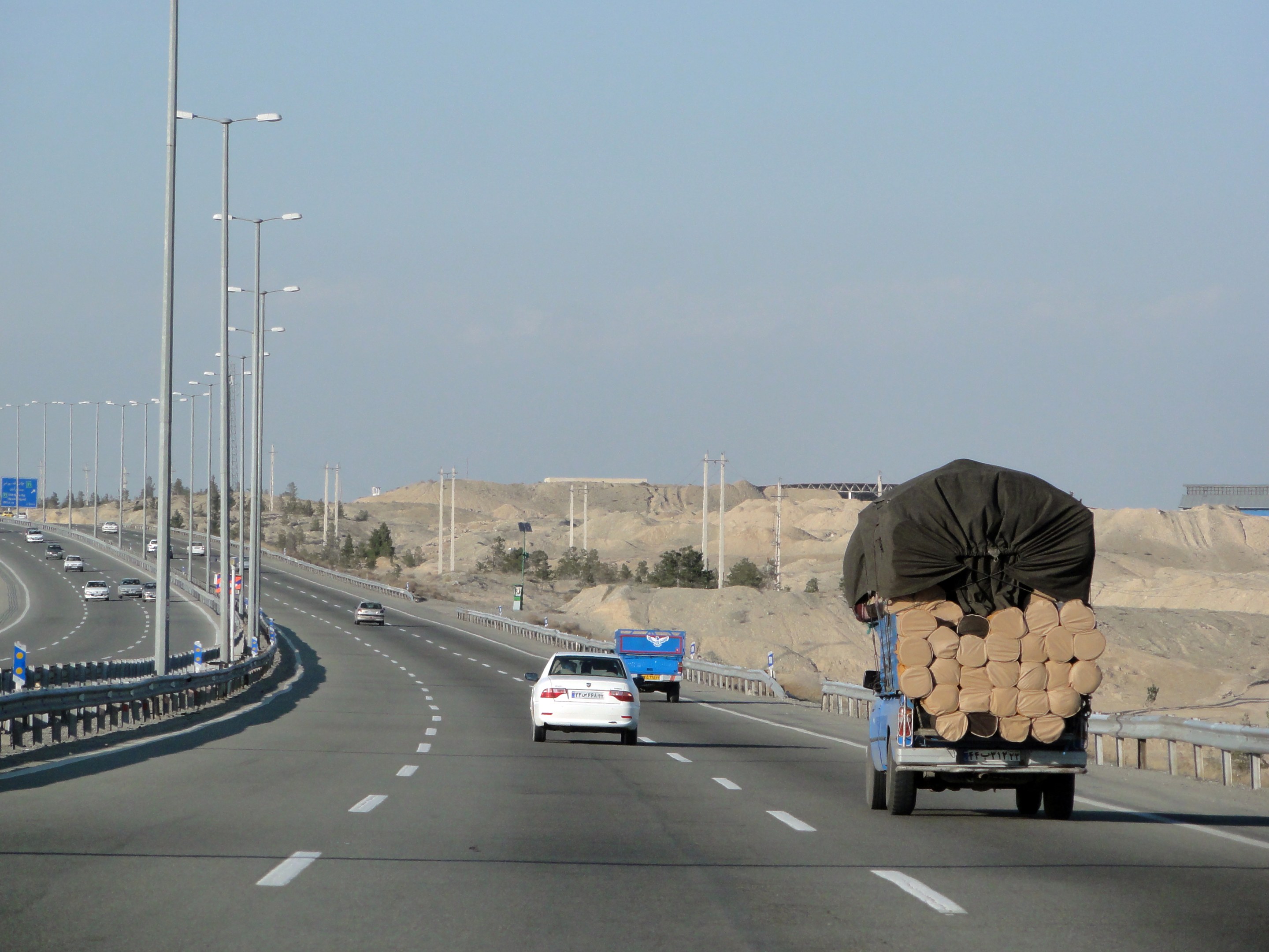 Ein Holztransporter mit einer großen Ladung Holz fährt auf einer Straße mit Leitplanken, Laternen, Schildern, Bäumen und Sand, mit Hügeln und einem klaren blauen Himmel im Hintergrund.