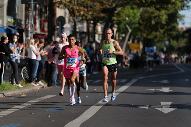 Gruppe von Läufern bei einem Marathon auf einer Stadtstraße mit Zuschauern, Grünflächen und städtischen Elementen im Hintergrund.