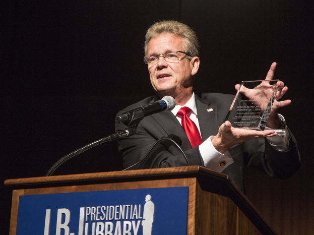 Ein Mann im Anzug und mit Krawatte steht an einem Rednerpult und hält eine Trophäe in der Hand, mit einem Mikrofon und einer Tafel daran, trägt eine Brille vor einem dunklen Hintergrund auf einer Bibliothekspreisverleihung.