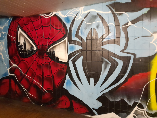 Ein buntes Wandgemälde von Spider-Man in einem roten und blauen Kostüm mit einem Webmuster und einem Spinnenlogo, dargestellt auf der Seite eines Gebäudes mit einem Boden darunter und einer Decke darüber.