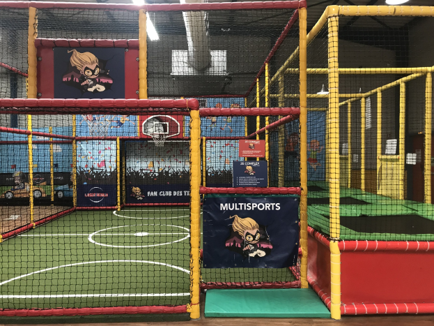 Großes Indoor-Trampolinpark mit einem Basketballfeld in der Mitte, umgeben von einem Netz und Wandbannern, mit einem Basketballkorb, Deckenleuchten und sichtbarem Boden.