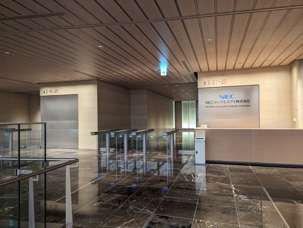 Eingangshalle eines Gebäudes mit Marmorboden, Glasgeländern, elektrischen Leuchten an der Decke, Namensschildern an den Wänden und einem Empfangstisch mit dem NEC Network & System Integration Corporation-Logo.
