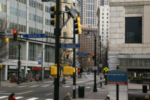 Eine belebte Stadtstraße mit hohen Gebäuden, Fußgängern auf dem Gehweg, Fahrzeugen auf der Straße, Verkehrsampeln und einem Baum im Vordergrund.