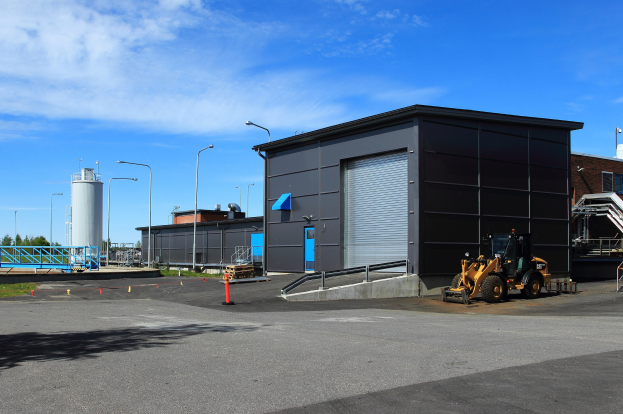 Großes Gebäude mit einem Bagger davor, umgeben von Straßeninfrastruktur und Fahrzeugen, unter einem bewölkten Himmel, als die neue Einrichtung im Industriepark bezeichnet.