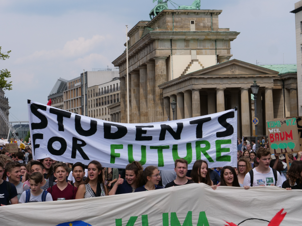 Eine Gruppe von Sch├╝lern marschiert in Berlin, mit einer bunten Fahne, auf der 'Students for Future' steht, vor dem Hintergrund von Geb├Ąuden, B├Ąumen und Himmel.