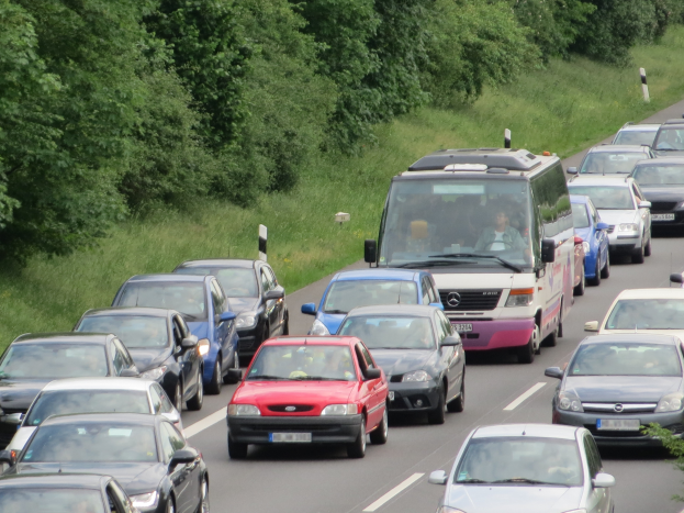 Ein Stau auf einer Autobahn mit vielen Autos und einem Van, Menschen in den Fahrzeugen sichtbar, mit Bäumen und Gras im Hintergrund.