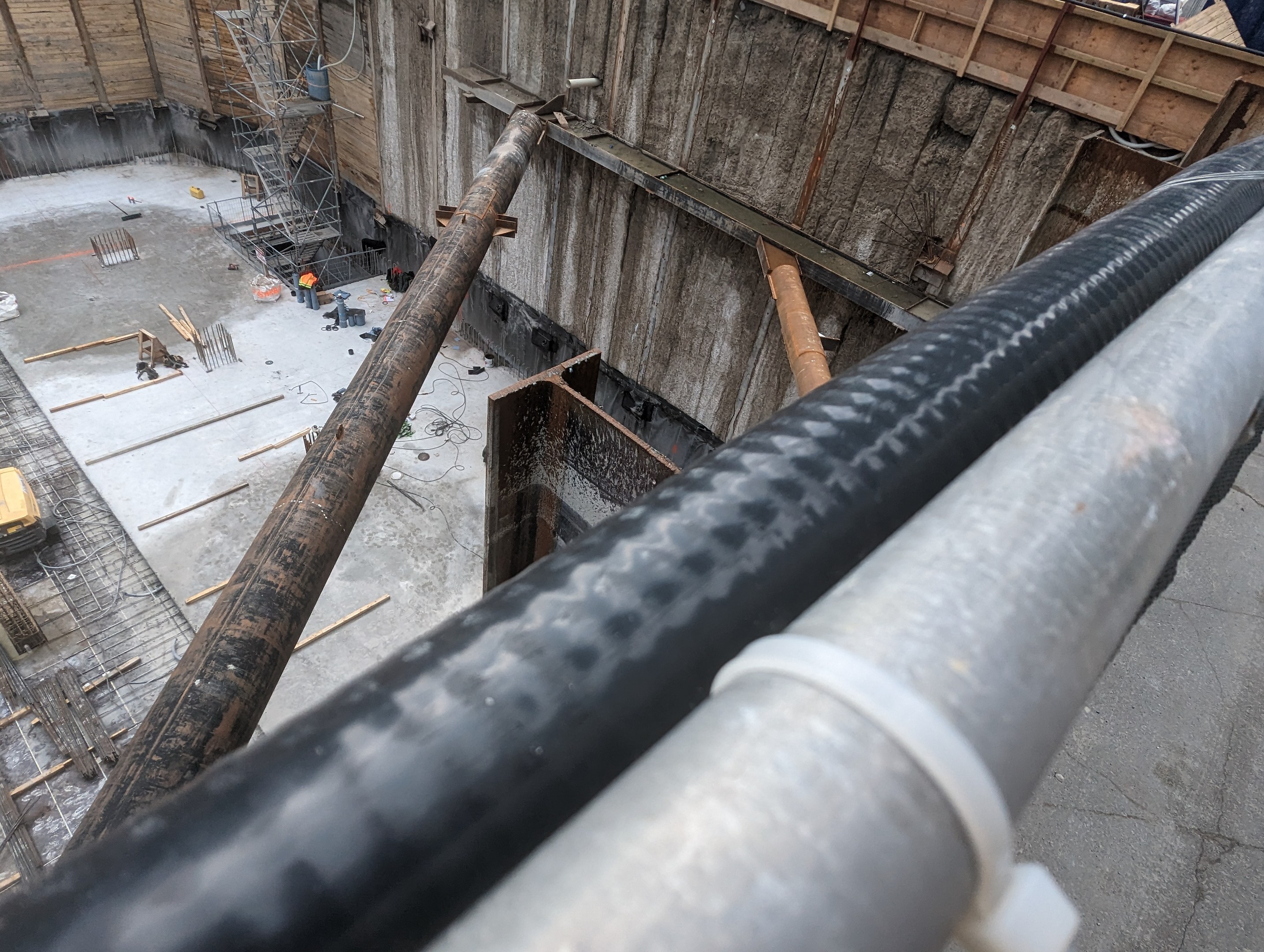 Großes Metallrohr auf einer Baustelle umgeben von Menschen, Holzgegenständen und anderen Materialien, mit einer Person in einem blauen Hemd rechts und Fahrzeugen und einem Gebäude im Hintergrund.