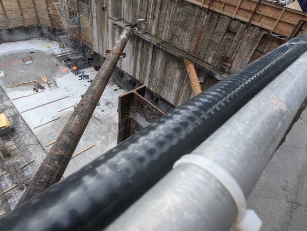 Großes Metallrohr auf einer Baustelle umgeben von Menschen, Holzgegenständen und anderen Materialien, mit einer Person in einem blauen Hemd rechts und Fahrzeugen und einem Gebäude im Hintergrund.