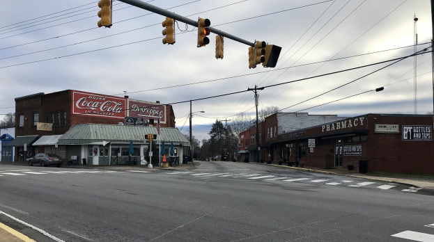 Stadtstraße mit Ampel an einer Kreuzung, Fahrzeuge, Gebäude mit Namensschildern, Strommasten mit Drähten, Bäume und ein bewölkter Himmel im Hintergrund.