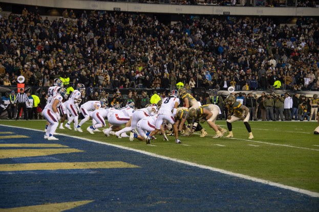 Fußballspieler in Sportkleidung und Helmen auf dem Boden während eines Spiels in einem Stadion mit Zuschauern und Beschilderung im Hintergrund.
