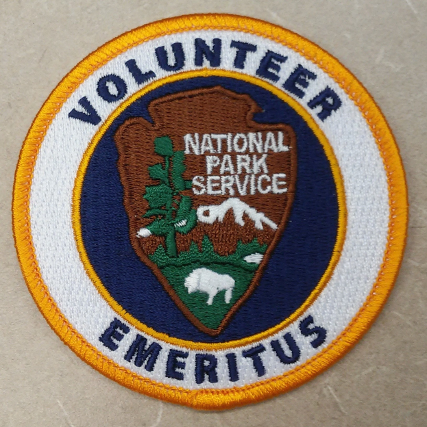 Runder Aufnäher mit blauem Rand und weißem Hintergrund, mittig die schwarze Schrift "Volunteer National Park Service Emeritus" in einem gelben Kreis.
