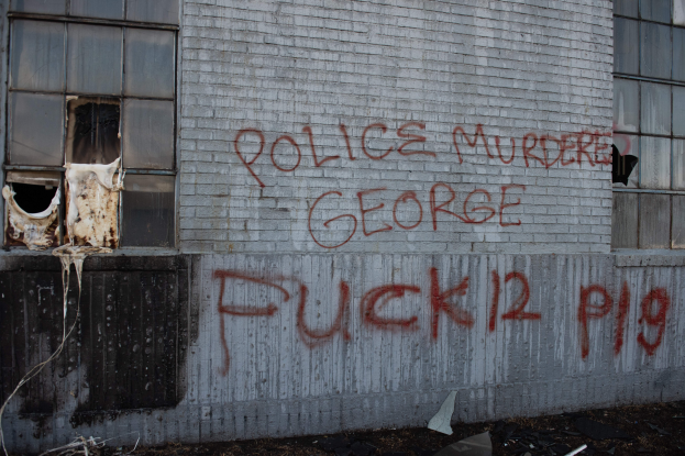 Ein Gebäude mit Graffiti, das "Polizei Mord George Fuck 12 P19" auf der Seite trägt, mit Fenstern und verschiedenen Gegenständen vorne.