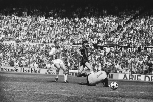 Schwarzes und weißes Foto von Männern, die auf einem Feld Fußball spielen, mit Zuschauern im Hintergrund und einer Fahne mit der Aufschrift "Bundesliga 1978-79 Ajax 1-1 Hannover 96."