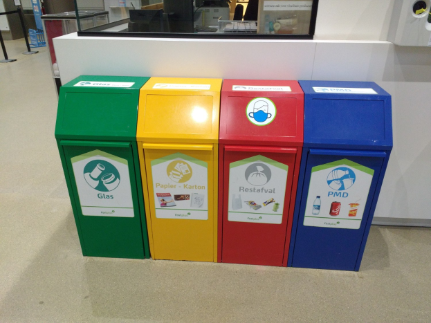 Vier bunte Mülltonnen in einer Reihe auf dem Boden, mit einer Arbeitsplatte, einem Monitor und anderen Gegenständen im Hintergrund und einer Person auf der linken Seite.