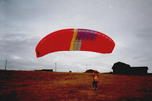 Eine Person mit einem roten Fallschirm in einem Feld, umgeben von Häusern, Pfählen und einem klaren blauen Himmel.
