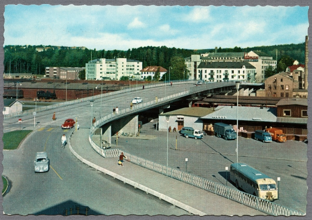 Altes Schwarz-Weiß-Foto einer Stadtstraße mit Autos, Bussen, Fußgängern auf einer Brücke, Laternenpfählen, Gebäuden mit Fenstern, Bäumen und einem bewölkten Himmel.