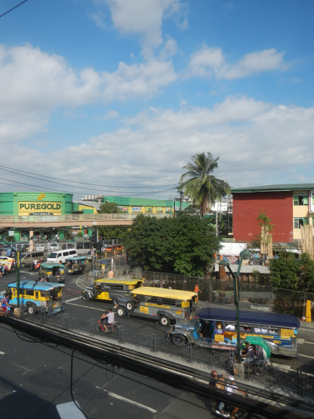 Eine belebte Stadtstraße mit verschiedenen Fahrzeugen, Fußgängern auf Gehwegen, Gebäuden, Bäumen, einem Zaun, Strommasten mit Drähten und einem bewölkten Himmel.